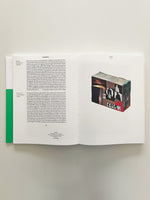 An overhead view of an open art book on a white surface. The left page has columns of text in English and German next to a vertical green bar. The right page displays a photograph of a rectangular sculpture covered in a dense collage of images and textures.