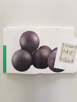 A top-down photo of an open art book on a plain surface. The book displays a photograph of several large, dark, spherical sculptures with Arabic script carved into them. A small card for the poetry book from A to Z by Etel Adnan rests on the right-hand page.