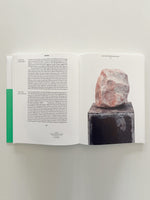An open book lies flat on a white surface. The left page contains two columns of text next to a vertical green bar. The right page features a large photograph of a rough, pink and white stone sculpture on a dark, textured pedestal.