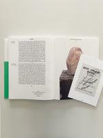 An overhead view of an open book laid flat on a white surface. The book displays text in English and German alongside a photograph of a pink, torso-like stone sculpture. A small white card with a map of Lebanon and the text Sitt Marie Rose by Etel Adnan is placed on the right page.