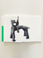 An overhead view of an open art book on a plain, off-white surface. The pages show a single photograph of a dark, abstract sculpture with a rough texture, resembling a figure riding a four-legged animal. The text Warrior, 2020 is printed on the top right. A bright green strip is visible on the far left edge of the book.