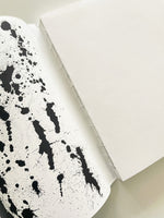 A high-angle, close-up shot of an open notebook with an exposed stitch binding. The left-hand inside cover is white and covered with artistic black ink splatters, which stands in stark contrast to the clean, blank white pages on the right.