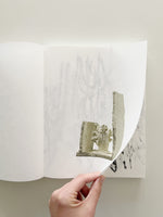 An overhead view of a hand turning the right page of an open art book. The page has a photograph of a small, beige sculpture resembling a plant fragment. As the page curls, it reveals a faint charcoal drawing on the sheet underneath.