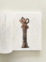 An open book lies on a plain, off-white surface. The right page displays a photograph of a tall, rust-colored, and dark-textured metal sculpture with a solid base and an abstract, looped top. Faint, gray, vertical brushstrokes decorate the background of both pages.