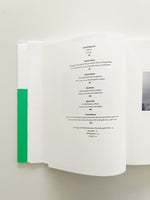 A high-angle shot of an open book lying on a flat surface. The right page displays the table of contents, with text in a black serif font listing authors and titles in both English and German. A bright green vertical stripe from the book's binding or cover is visible on the left.