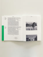 A flat lay photograph of an open book spread. The left page has a green vertical bar on its edge and columns of text in both English and German. The right page features two black and white photographs stacked vertically: the top one is a portrait of a smiling woman on a balcony, and the bottom one is a view of a harbor with buildings and a ship.