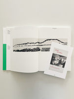 A top-down view of an open art book with a green spine, laid on a light surface. The pages display a wide, minimalist landscape created with black ink brushstrokes. Resting on the right page is a smaller booklet titled RŪMĪ and Sufism, featuring a black-and-white photo of whirling dervishes on its cover.