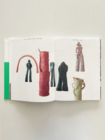 A top-down view of an open art book laid flat on a white surface. The two-page spread displays photographs of several colorful, abstract, and figurative clay sculptures against a stark white background. The sculptures vary in shape and color, including red, dark green, and muted tones. The book's spine, with a bright green design element, is visible on the left.