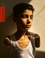A powerful bust portrait of a young boy who is a double amputee, wearing a white mesh tank top. Strong sunlight from the side illuminates half of his pensive face and body against a plain wall, casting the other half in deep shadow. The image is a book cover for World Press Photo 2025.
