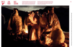 A group of Chinese migrants in hooded plastic ponchos huddle together for warmth around a fire at night. The somber scene at the US-Mexico border is illuminated only by the warm, orange glow of the flames from below, casting long shadows in the darkness.