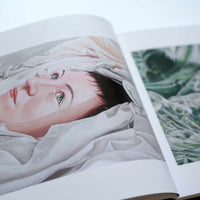 An angled, close-up photograph of an open art book. The left page features a hyperrealistic painting of a person with fair skin, short dark hair, and vivid green eyes lying partially submerged in water amongst folds of white fabric.