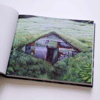 An open art book lying on a white surface, displaying a detailed painting of a rustic shed with a turf roof. The shed, made of patchwork metal and wood, is nestled into a lush green hillside and is being overgrown by tall grasses and foliage. A small, white owl is painted on the side of the building.