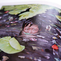 An angled, close-up photograph of artwork printed in a book. The image shows a young boy with dark hair submerged to his nose in murky, polluted water. He is squinting his eyes in a smile, surrounded by floating garbage and a few green lily pads.