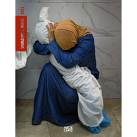 A woman in a blue dress and tan headscarf kneels on a tiled floor, her head bowed in grief as she cradles a body wrapped in a white shroud.