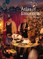 The book cover for Atlas of Emotions by Jari Silomäki shows a chaotic party scene where several cats are on a table covered with cake and cookies, one wearing a tiny party hat, while people celebrate in the background.