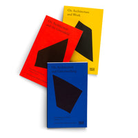 Three paperback books with solid blue, red, and yellow covers are fanned out against a white background. Each book features a large, abstract black shape and white text. The titles are On Architecture and Greenwashing, On Architecture and the Greenfield, and On Architecture and Work.