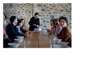 An interior shot shows a woman with long dark hair dressed in black, standing at the head of a long wooden table and serving food from a pot into a bowl. Several people are seated at the table, looking on in a communal dining setting in front of a rustic stone wall.