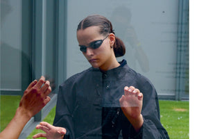 A young woman with her dark hair slicked back and wearing small, oval sunglasses and a dark jacket stands behind a pane of glass. She looks down with a serious expression as her hands and another person's hands are held up to the glass, with reflections visible on the surface.