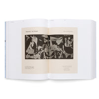 An angled photograph of an open book with a light blue cover, set against a white background. The book displays a two-page spread featuring a black-and-white reproduction of Pablo Picasso's cubist painting, Guernica. Text in both a Latin script and Arabic script accompanies the image.