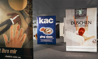 An interior shot of a gallery displaying several large, surreal posters with German text. From left to right, a poster shows a headless body and a glowing hand, a blue poster features white sausages, and a third shows a woman in a bathtub holding up a television with her feet.