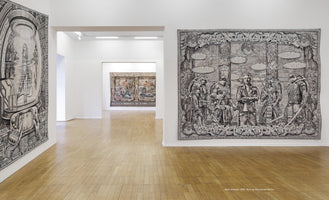 An interior view of a modern art gallery with white walls and light hardwood floors. On display are several large, intricate tapestries. In the foreground, a black-and-white tapestry on the right wall features four characters resembling the Teenage Mutant Ninja Turtles flanking a man. Another tapestry is partially visible on the left, and a more colorful, classical-style tapestry hangs at the end of a distant hallway.