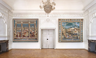 An ornate, white-walled room with a high ceiling, a crystal chandelier, and a parquet floor. Two large, colorful, and detailed tapestries hang on the main wall, flanking a central white double door. The room is also furnished with two marble fireplaces.