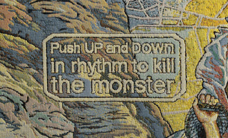 A close-up of a woven tapestry depicting a video game instruction. In a text box with a rounded border, the words Push UP and DOWN in rhythm to kill the monster are woven in a blocky, pixel-like font against a light blue background. The surrounding tapestry features a rocky landscape in shades of blue, yellow, and brown.