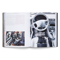 An open art book displayed on a white surface. The right page shows a full-page photograph of a car's interior, with the steering wheel, dashboard, and driver's seat covered in thick, expressive strokes of black and white paint. The left page contains text in German and English, and a smaller photo of the painted car with its doors open.