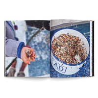 An open book showing a two-page photo spread. On the left page, a child wearing a winter coat holds out a handful of birdseed; their fingernails are painted blue. On the right page is a close-up of a large white and blue enamel pot full of a mix of birdseed, nuts, and corn.