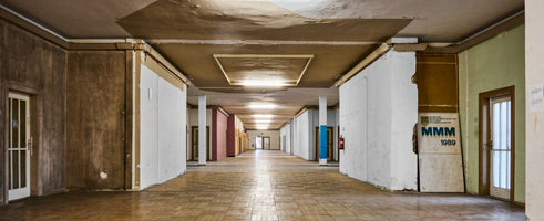 A wide, symmetrical photograph of a long, dilapidated hallway in an abandoned building. The ceiling paint is peeling, walls are a patchwork of bare, white, and wood-paneled sections, and the tiled floor stretches into the distance. A sign on the right reads MMM 1989.