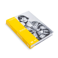 An angled view of the hardcover book Coco Chanel's Roaring Twenties against a white background. The cover features a black-and-white photograph of Coco Chanel, wrapped in a prominent, bright yellow band that shows the title in white lettering.