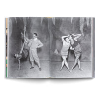 An open book displays a vintage black-and-white photograph across two pages. On the left, a man in a striped sweater helps a woman in a leotard with a one-handed handstand. On the right, two women in knit athletic wear pose closely, one whispering to the other in front of a painted backdrop.