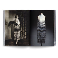 An open book displays a two-page spread about a 1920s flapper dress. The left page shows a vintage black-and-white photograph of a woman wearing the heavily beaded dress, complete with pearls and a feather fan. The right page features a modern, full-color photograph of the same dress on a mannequin against a dark background, highlighting the intricate, shimmering beadwork that creates an ombré effect from white to black.