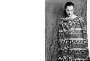 A black and white portrait of a person with dramatic, pale makeup featuring sharp winged eyeliner and a painted widow's peak. Their dark hair is slicked back, and they stare intensely at the camera. They wear a voluminous, off-the-shoulder poncho with horizontal bands of a geometric, step-like pattern against a dark, wrinkled backdrop.