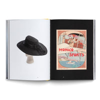 An overhead photograph of an open book laid flat. The left page has a white background and shows a black, wide-brimmed hat with a ribbon on a mannequin head. The right page has a black background and displays a vintage-style illustrated poster for Monaco Sports, featuring a woman in a hat driving a boat.