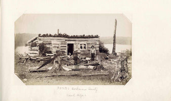 An old, sepia-toned photograph of four people and a dog posing outside a rustic log cabin by a lake. The group includes three women in long dresses and a man in a hat and suspenders. The foreground is littered with large tree stumps and a two-man saw, indicating a logging area.