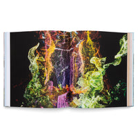 An open book on a white surface displays a vivid, abstract image across a two-page spread. Against a black background, granular, flame-like shapes in bright green, yellow, orange, and purple swirl upwards from the center of the pages.