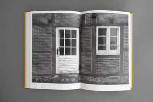 An open book with a yellow hardcover lies flat on a grey surface, displaying a full-spread, black-and-white photograph of a rustic brick and timber-framed house with a weathered white door and a matching window.