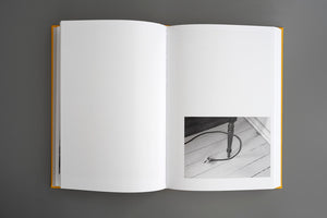 An overhead view of an open book with a yellow cover on a gray background. The pages are blank, except for a small black-and-white photo on the bottom right showing an unplugged electrical cord and a table leg on a white wood floor.