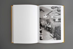 An open, yellow-spined hardcover book lies flat on a gray surface. The left page is blank, while the right page features a black and white photograph of the interior of a cluttered shop. A life-sized statue of a horse stands in the foreground, looking into the room filled with various objects, furniture, and curiosities.