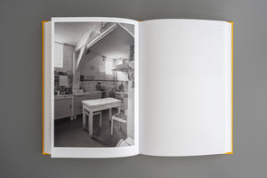 An open, yellow-covered book laid flat on a grey surface. The left page features a black-and-white photograph of a dated, utilitarian kitchen with tiled walls and a small table. The right page is blank.