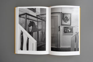 A black and white photograph spread across the two pages of an open book with a yellow cover. The photo shows an interior scene from a staircase landing. On the left page, a wooden horse head is visible inside a glass display case. On the right page, a wall has a door and two framed pictures.