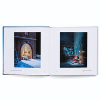 An open art book displaying two photographs of detailed miniature scenes. The left-hand page shows a snowy exterior at night, with a view through an arched window into a warm, lit dining room. The right-hand page shows a dimly lit, wood-paneled library with a red armchair sitting in a pool of light.