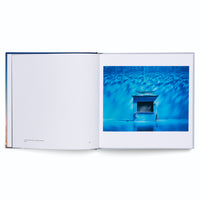 An open photography book on a white surface. The right page features a surreal photograph of a closed kiosk labeled TRINKHALLE. The entire scene is saturated in a deep blue color, with dappled light patterns on a tiled wall and a reflective, wet-looking floor, giving the impression of an underwater setting. The left page is mostly blank with a small caption.