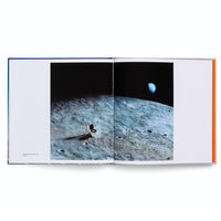 An open book on a white surface displays a surreal two-page photograph. The image shows a person sitting on a white toilet placed on the gray, cratered surface of the moon. In the blackness of space behind them, the blue and white planet Earth is visible in the distance.