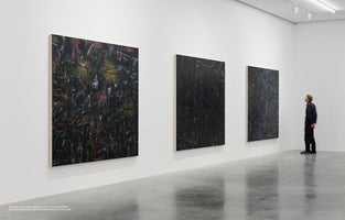 A wide shot of a modern art gallery with white walls and a polished concrete floor. Three large, dark, vertical abstract paintings covered in frenetic, light-colored scribbles hang in a row. To the far right, a person in dark clothing stands looking up at the artwork.