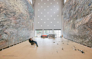 A wide-angle view of a massive, high-ceilinged artist studio. On the left and right, two monumental, wall-sized abstract paintings are filled with a dense, energetic storm of black lines and marks. In the foreground, artist Julie Mehretu sits in a lounge chair, dwarfed by her creations as she contemplates her work.