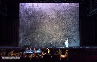 A wide view of a theater stage during a performance of the opera Only the Sound Remains. In the foreground orchestra pit, musicians and a choir perform. On stage, a dancer in a white, flowing costume poses in front of an immense, abstract backdrop by artist Julie Mehretu, featuring energetic, dark brushstrokes on a light gray background.
