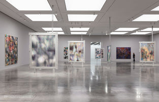 A wide interior view of a contemporary art gallery with a polished grey floor and white walls. Large, abstract paintings are displayed; some colorful graffiti-style pieces hang on the walls, while several semi-translucent, blurry paintings in heavy white frames are suspended from the ceiling. A lone visitor stands in the background, viewing the art.