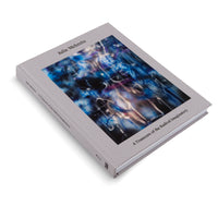 A light gray hardcover book titled Julie Mehretu: A Transcore of the Radical Imaginatory, displayed at an angle on a white background. The cover features a vibrant, abstract artwork with energetic, swirling lines of blue, purple, and glowing white light against a dark, blurry backdrop.